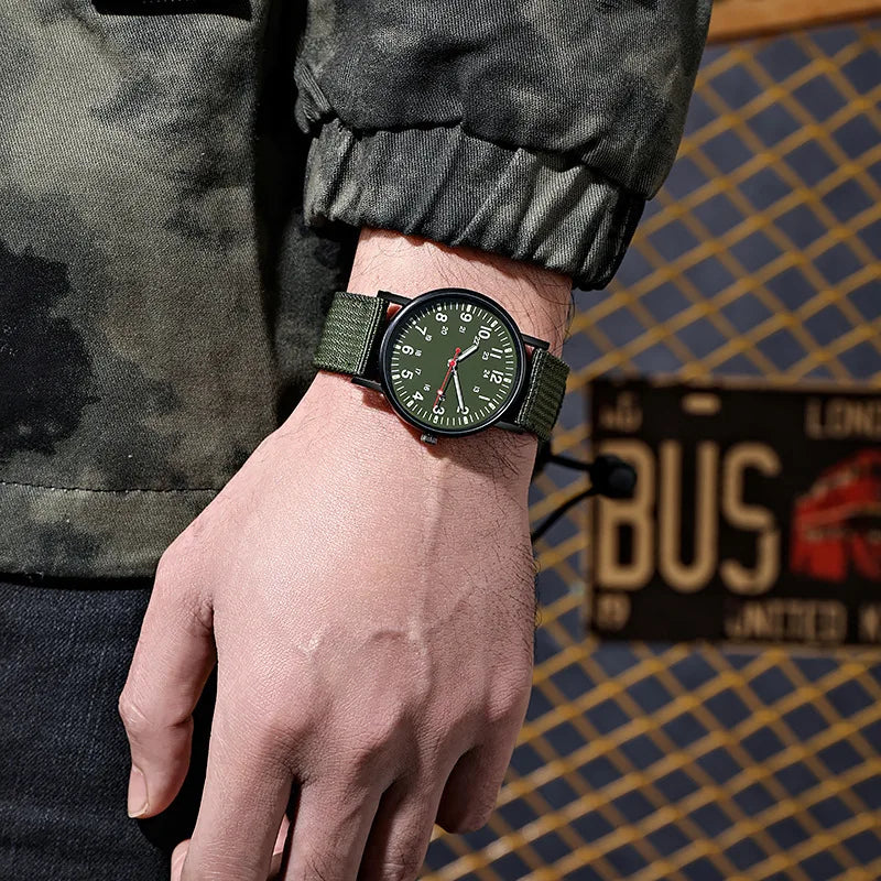 Military field watch in green worn on a man's wrist with camo sleeve, outdoor lifestyle shot