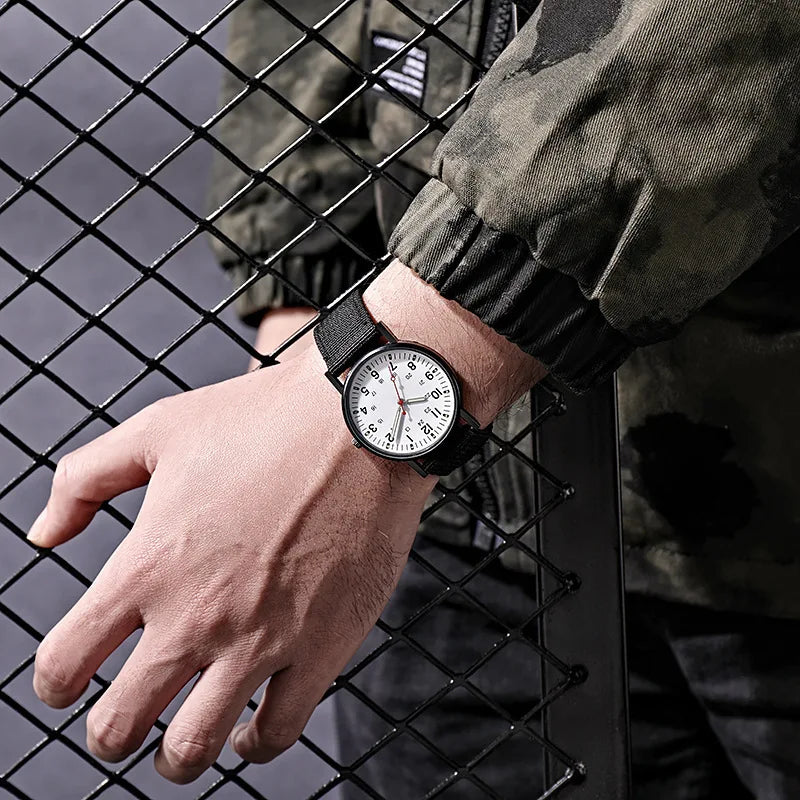 Military field watch with white dial and black strap on wrist, hand gripping a chain-link fence outdoors