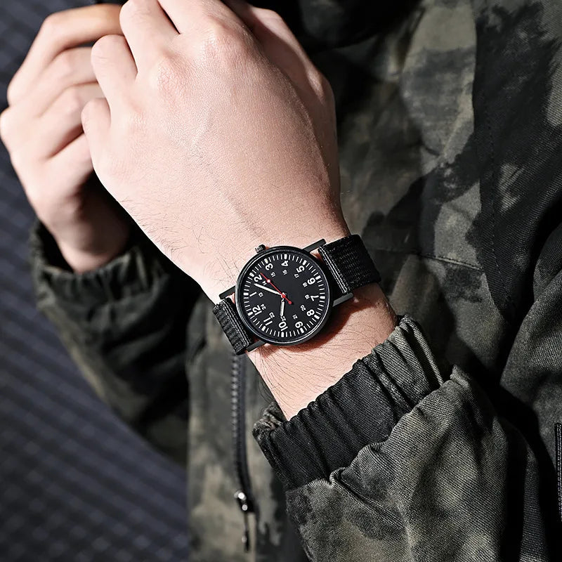 Military field watch in black with nylon strap worn on wrist, close-up lifestyle shot with camo jacket
