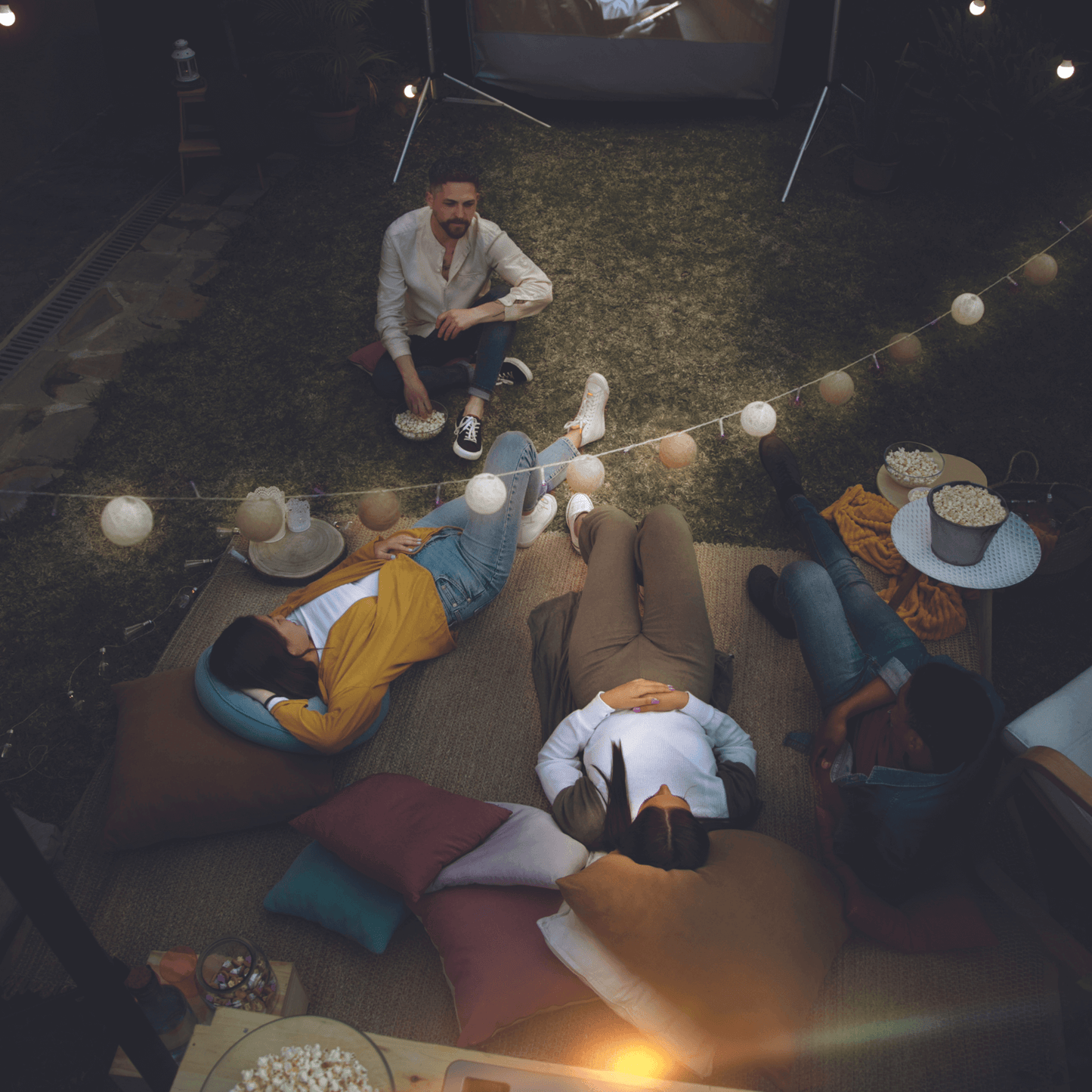 Group of people sitting on bean bags under string lights at night watching a movie from portable projector tech gift for dad who loves movies. 