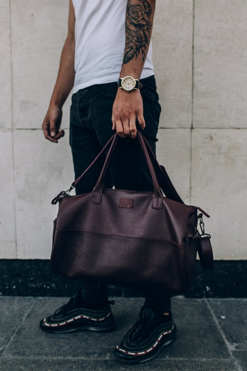 Men's genuine leather gym duffel sports bag — fitness gift for active dad A person wearing a white sleeveless top, black pants, and black sneakers carries a large dark brown leather duffel bag against a light stone wall.