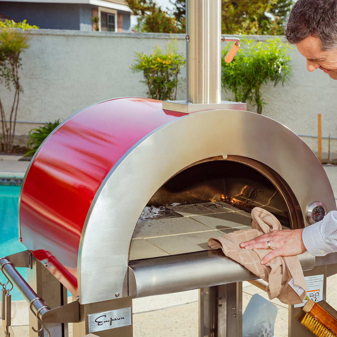 Person using a red and silver outdoor pizza oven with a pool and garden in the background. Empava outdoor wood-fired pizza oven — unique BBQ and cooking gift for dad